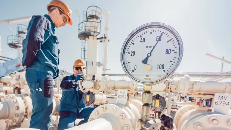 Two workers inspecting gas pipeline system with pressure gauge at industrial facility.